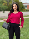 Miik model Yasmine (5'0", xsmall, petite) smiling wearing Miik's Janette puff sleeve reversible blouse in red wine tucked in a black pant