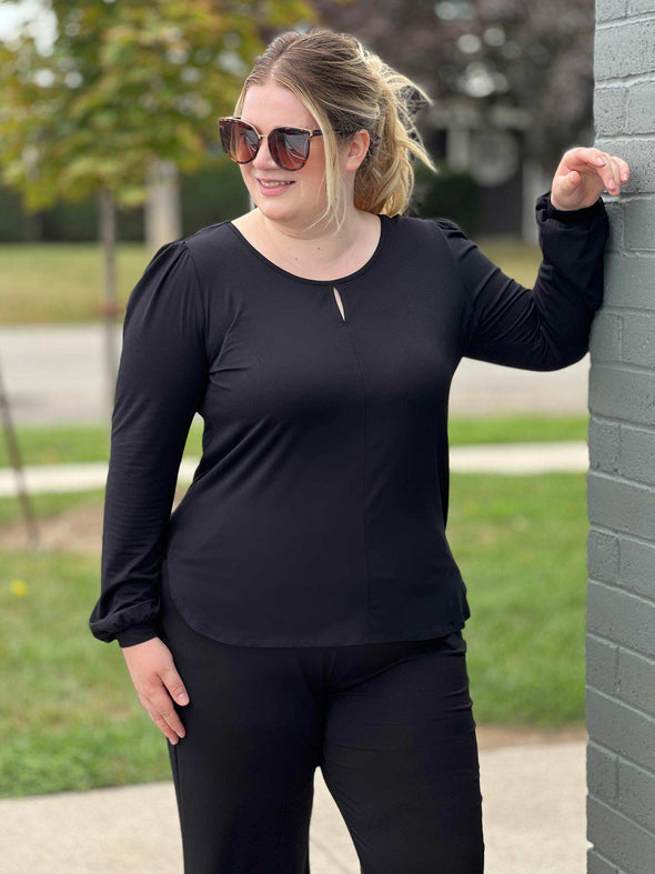 Woman standing next to brick wall looking away wearing Miik's Janette top in black with a matching colour pants and sunglasses