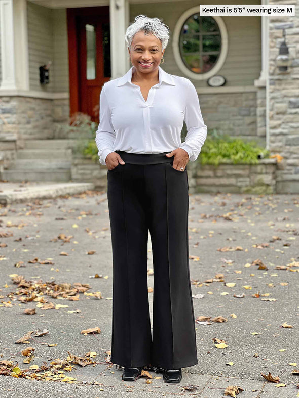 Miik model Keethai (5'5", medium) smiling with hands on pockets wearing Miik's Jeremy high waisted wide leg ponte pant in black with a collared white shirt tucked in