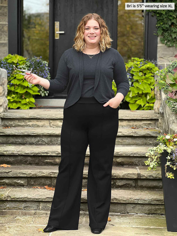 Miik model Bri (5'5", xlarge) smiling in front of a front door stairs wearing Miik's Jeremy high waisted wide leg ponte pant in black with a tank and cropped cardigan in charcoal 