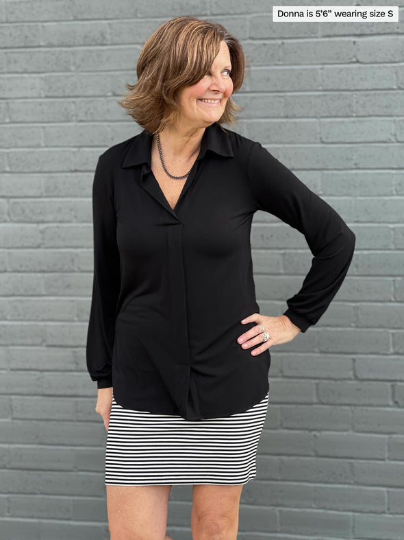Woman standing in front of a wall wearing Miik's June simple skirt/scarf in black and white stripe with a black collared shirt.