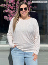 Miik model Christal (five feet three, large) smiling standing in front of a window wearing Miik's Kallyn slouchy dolman long sleeve top in cobblestone print with jeans