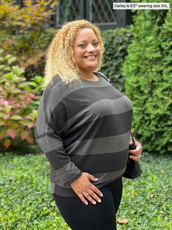 Miik model Carley (5'2", xxlarge) standing sideway smiling wearing Miik's Kallyn slouchy dolman long sleeve top in dark ash stripe and black legging