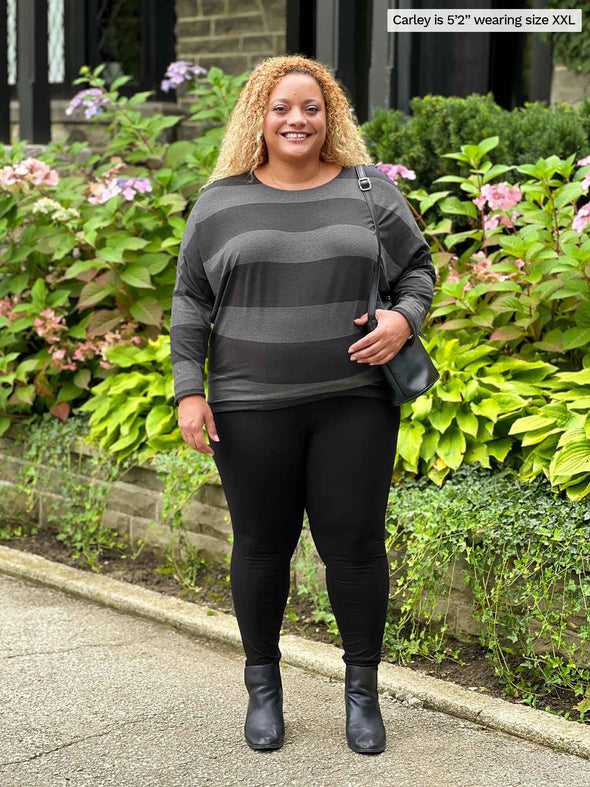 Miik model Carley (5'2", xxlarge) standing in front of a garden smiling wearing Miik's Kallyn slouchy dolman long sleeve top in dark ash stripe, black legging and boots