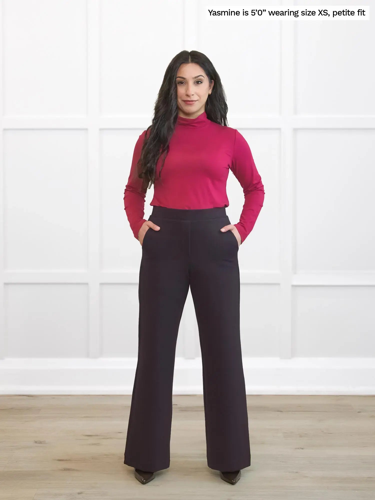Woman wearing a pink turtleneck and dark brown petite pants against a white background 