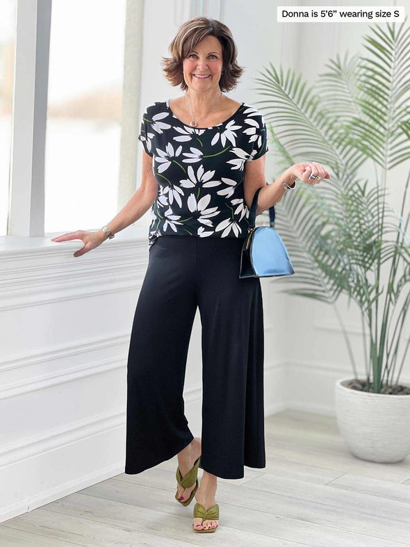 Miik founder Donna (5'6", small) smiling while standing in front of a window/white wall studio wearing Miik's Keethai wide leg culotte in black with a printed black/white flower tee