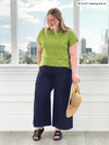 Miik model Bri (5'5", xlarge) smiling and looking down while standing in front of a window wearing a green moss top with Miik's Keethai wide leg culotte in navy 