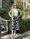 Miik model Liane (59", small) smiling while standing in front of a garden wearing Miik's Keethai wide leg culotte in white lily print, a white top and a machine washable blazer in green moss 