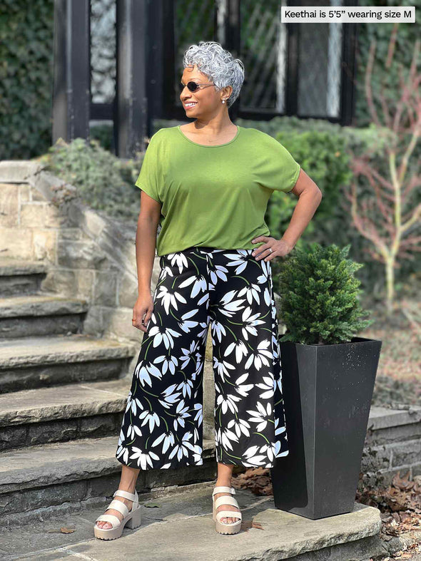 Miik model Keethai (5'5", medium) standing on a stairway looking away wearing a green moss top along with Miik's Keethai wide leg culotte in white lily print 
