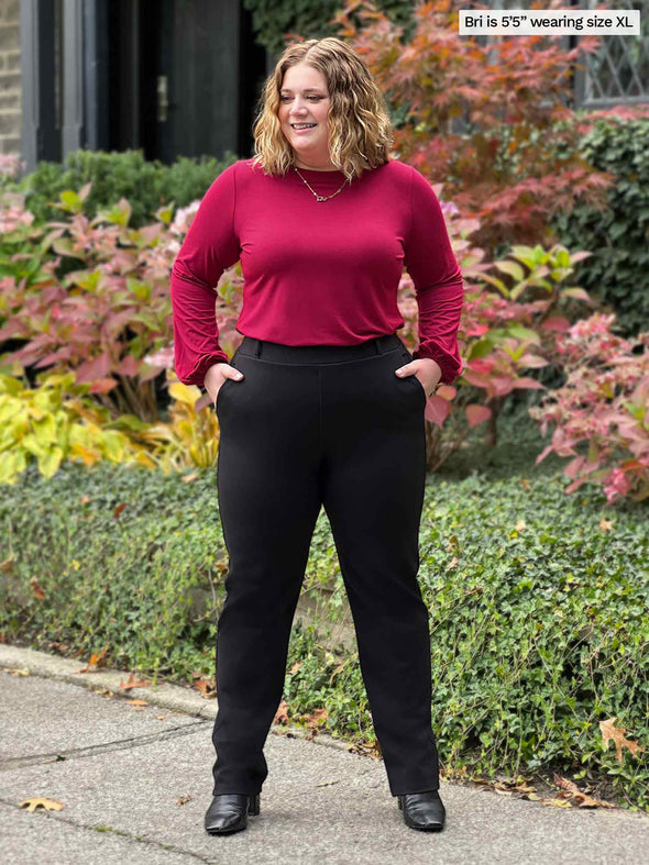 Miik model Bri (5'5", xlarge) smiling and looking away with hands on pockets wearing a bordeaux long sleeve top with Miik's Keltie ponte mid-rise slim pant in black