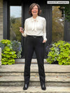 Miik founder Donna (5'6", small) smiling while standing on a front door wearing Miik's Keltie ponte mid-rise slim pant in black, with a belt in the same colour and a natural tie blouse