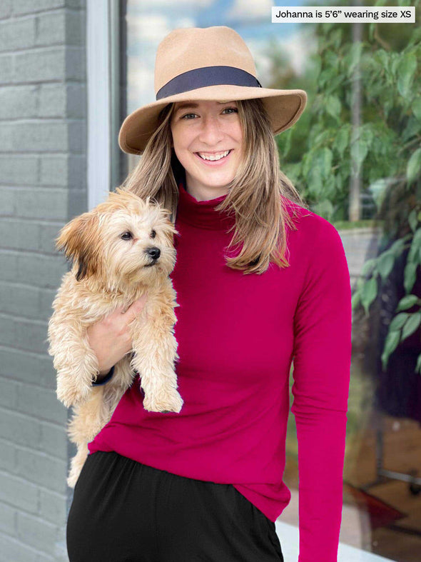 Miik model Jo (5'6", xsmall) smiling and holding a puppy wearing Miik's Kerry turtleneck top in bordeaux with a black pant and a hat 