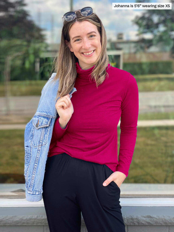 Miik model Jo (5'6, xsmall) smiling while standing in front of a window wearing Miik's Kerry turtleneck top in bordeaux with a black pant and a denim jacket over the shoulders 