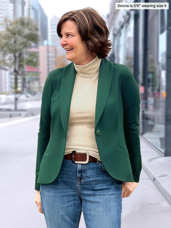 Miik founder Donna (5'6", small) smiling and looking away wearing Miik's Kerry turtleneck top in camel melange with jeans and a green pine blazer in a casual outfit 