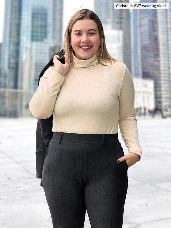 Miik model Christal (5'3", large) smiling while wearing Miik's Kerry turtleneck top in camel melange with a charcoal pinstripe pant and a matching blazer over the shoulders 