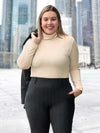 Miik model Christal (5'3", large) smiling while wearing Miik's Kerry turtleneck top in camel melange with a charcoal pinstripe pant and a matching blazer over the shoulders