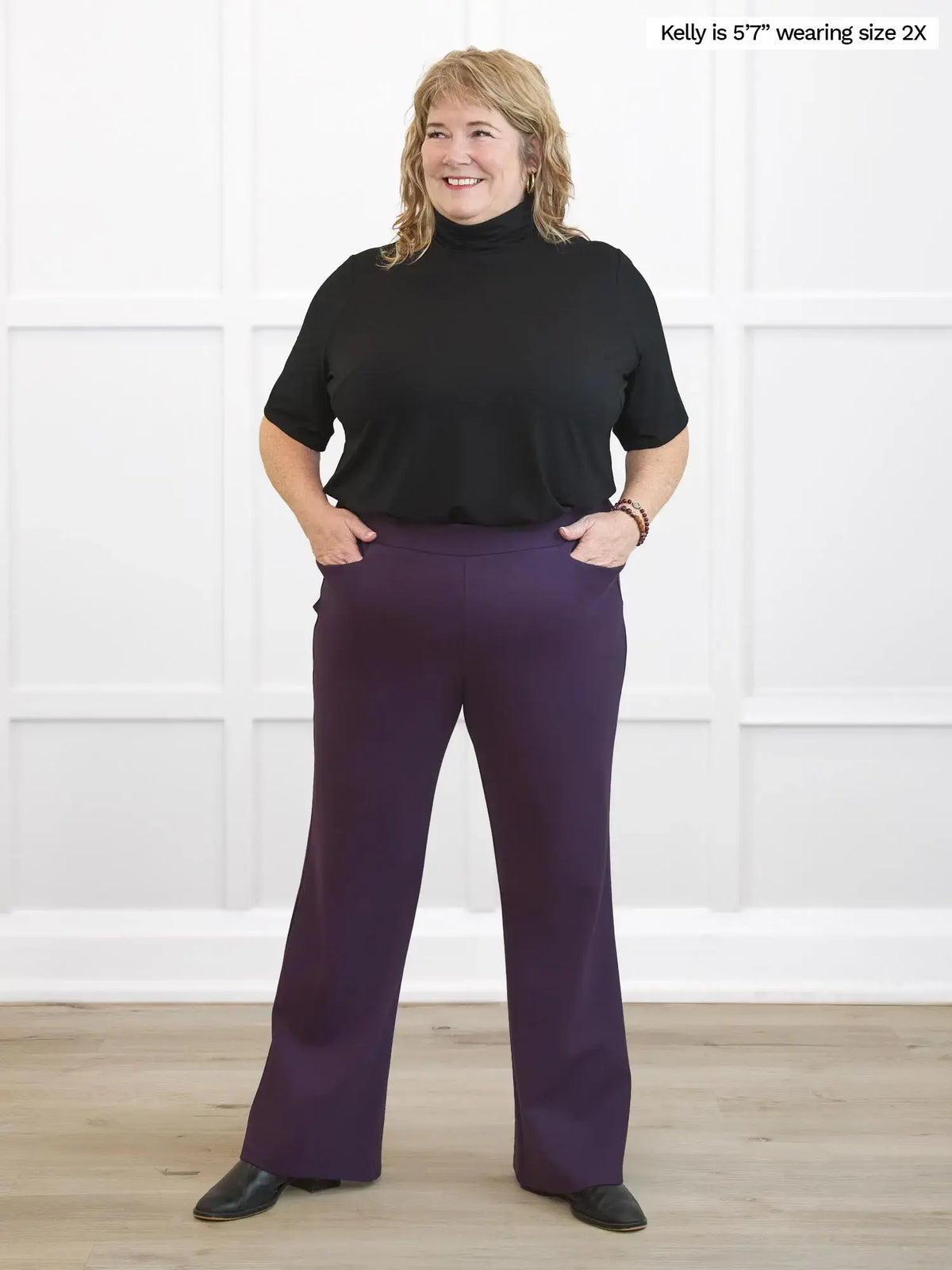 Woman wearing a black top and purple pants standing against a white wall. 