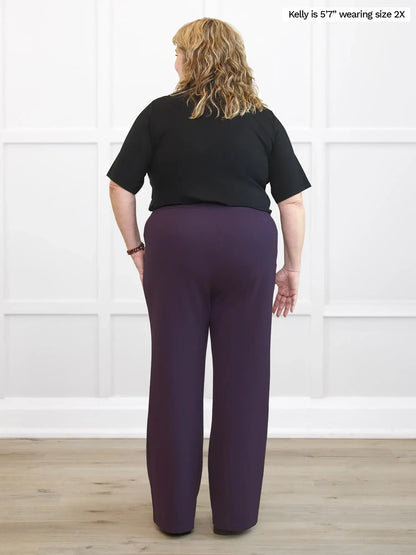 Woman wearing a black top and purple pants against a white wall. 