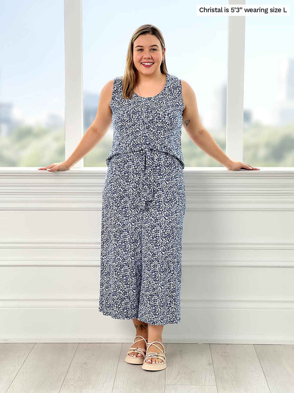 Miik model Christal (5'2", large) smiling with open arms while standing in front of a window wearing Miik's Kimmay open-back capri jumpsuit in baby's breath print 