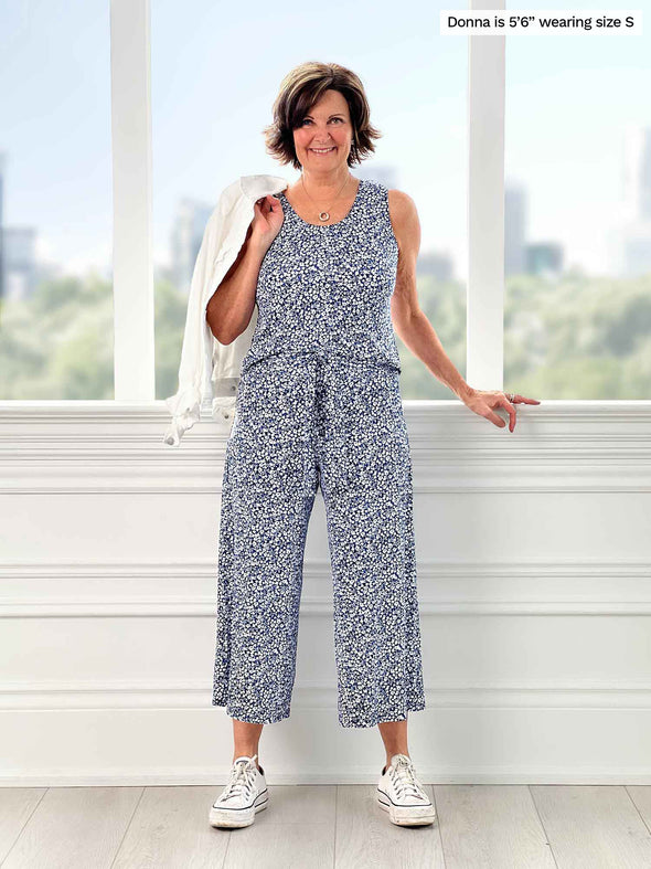 Miik founder Donna (5'6", small) smiling while standing in front of a window wearing Miik's Kimmay open-back capri jumpsuit in baby's breath print, white sneakers and a white denim jacket over her shoulders 