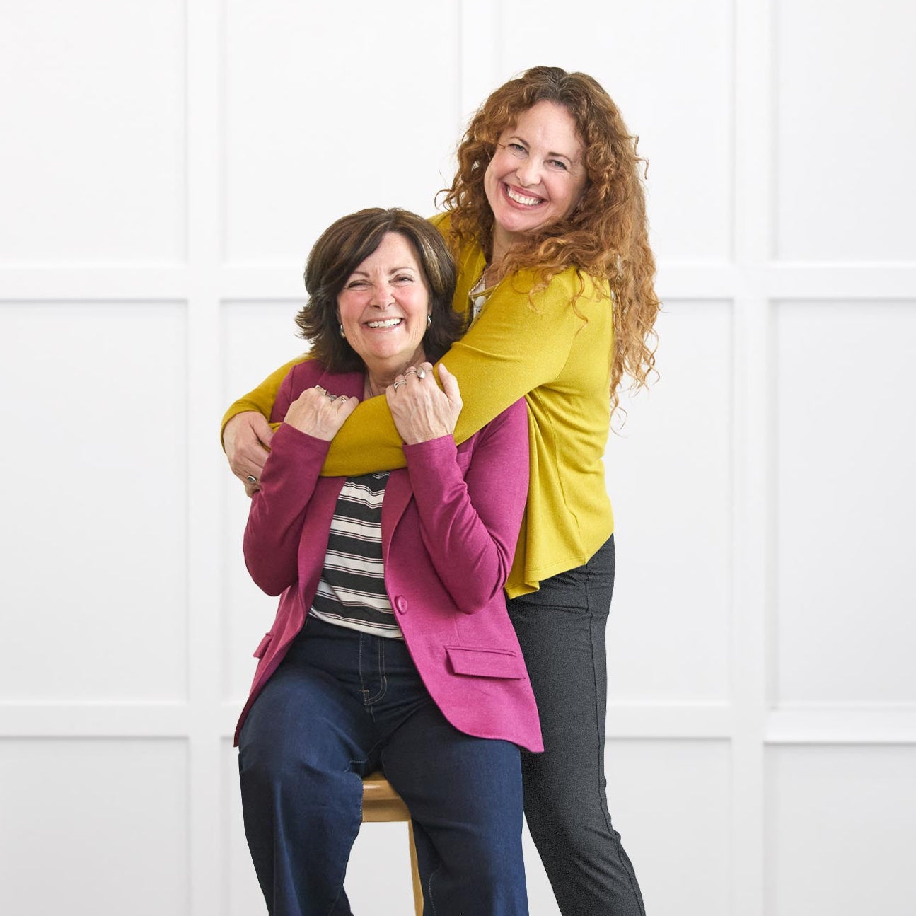 Two women posing together on a wooden floor with a white background