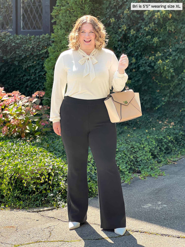 Miik model Bri (five feet five, xlarge) smiling in front of a garden wearing Miik's Miik's Laney mid-rise flare pant in graphite with a natural blouse tucked in