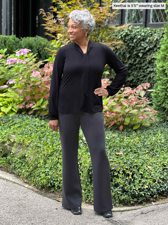 Miik model Keethai (five feet five, medium) smiling and looking away wearing Miik's Laney mid-rise flare pant in graphite and a black long sleeve tie shirt