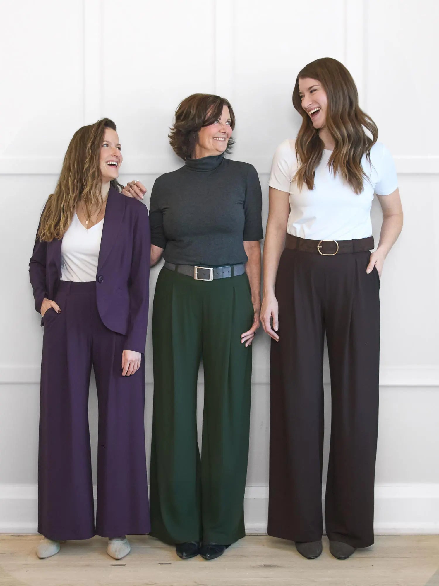 Three women standing together, each wearing different colored wide-leg pants against a white wall.