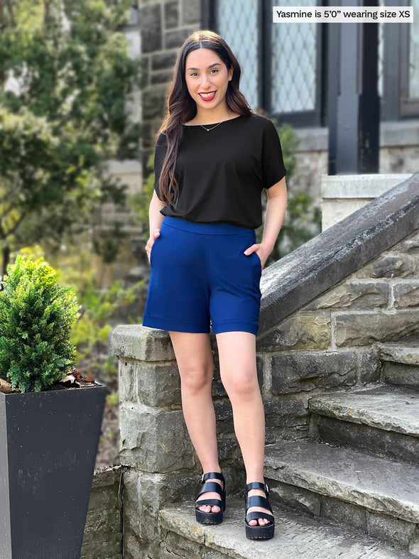 Miik model Yasmine (5'0", xsmall, petite) smiling while standing on a stairway wearing Miik's Leland everyday dressy short in ink blue with a black boatneck top 