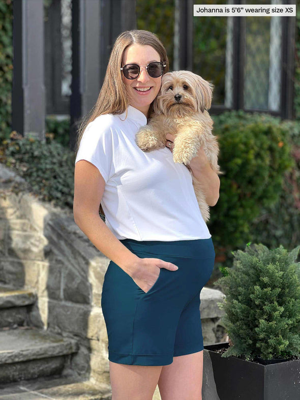 Miik model Jo (5'6", xsmall) smiling while standing sideway wearing Miik's Leland everyday dressy short in teal with a white shirt. Jo is also holding a puppy 