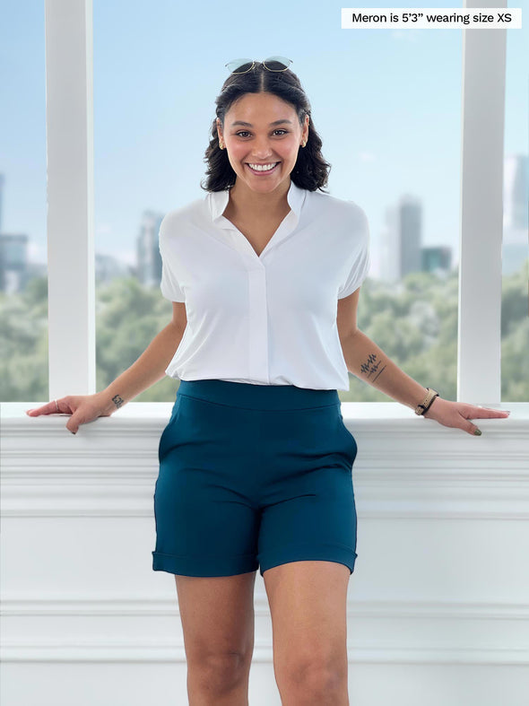 Miik model Meron (5'3", xsmall) smiling with open arms while standing in front of a window wearing a band collar top in white with Miik's Leland everyday dressy short in teal