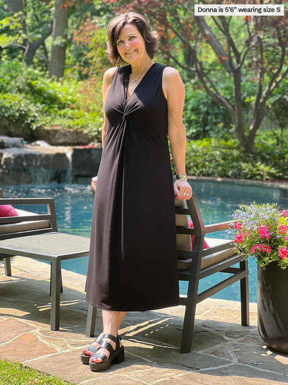 Miik founder Donna (five feet six, small) leaning against to a outdoor chair in front of a pool wearing Miik's Lina midi knot dress in black