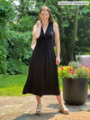 Miik model Johanna (five feet six, xsmall) standing on a backyard smiling wearing Miik's Lina midi knot dress in black holding a cardigan on her shoulders 