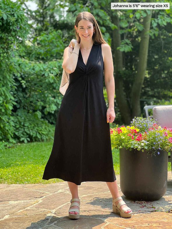 Miik model Johanna (five feet six, xsmall) standing on a backyard smiling wearing Miik's Lina midi knot dress in black holding a cardigan on her shoulders 