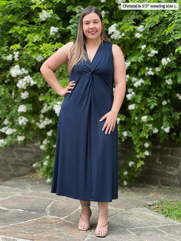 Miik model Christal (five feet three, large) standing in front of a garden smiling wearing Miik's Lina midi knot dress in navy 