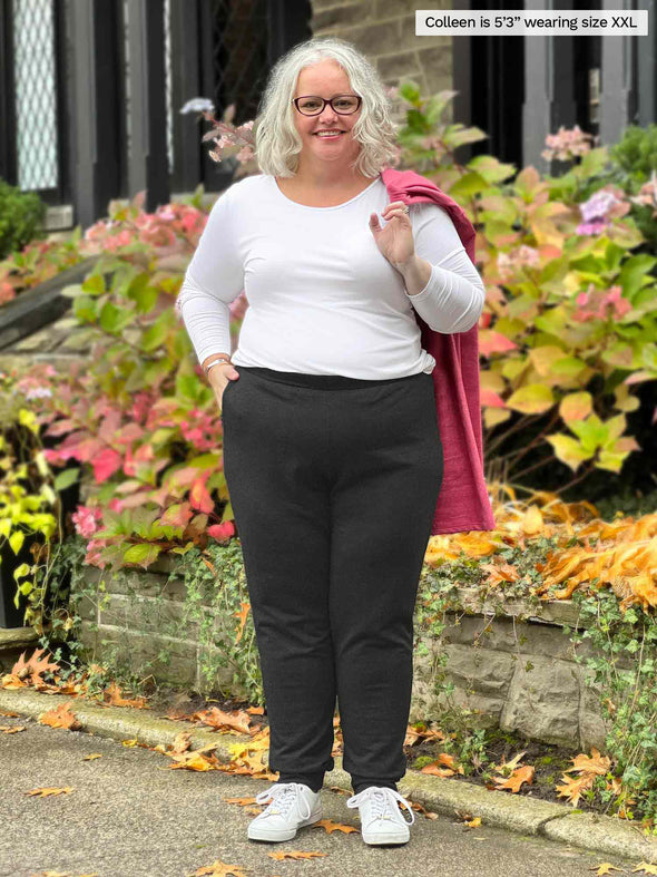 Miik model Colleen (5'3", xxlarge) smiling wearing Miik's Linaya luxe fleece jogger in charcoal with a long sleeve tee in white and a pink pomegranate cardigan over her shoulders
