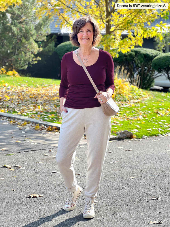 Miik founder Donna (5'6", small) smiling wearing Miik's Linaya luxe fleece jogger in oatmeal melange along with a long sleeve ruched top in port, a crossbody beige bag and white converse 