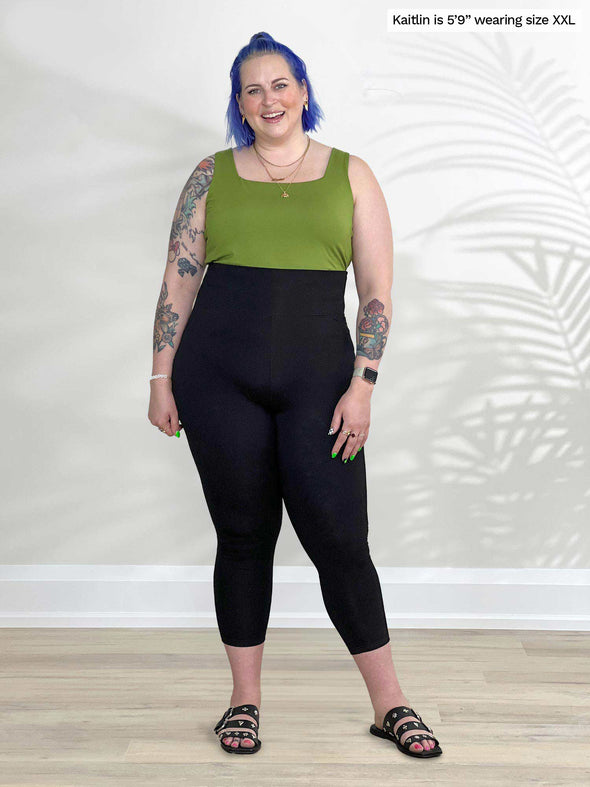 Miik model Kaitlin (5'9", xxlarge) smiling while standing in front of a white wall wearing Miik's Lisa2 high waisted capri legging in black with a green moss tank top 