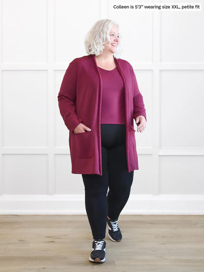 Woman wearing a long burgundy cardigan over a black outfit in a room with white walls. 