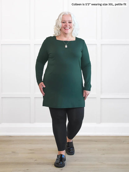 Woman wearing a green dress and black leggings standing against a white wall. 