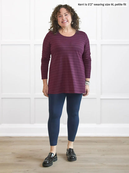 Woman wearing a purple striped long-sleeve top and blue leggings standing in a room with white walls. 