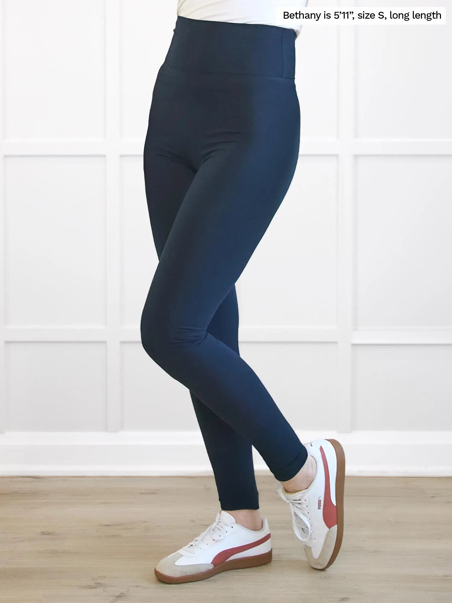 Person wearing navy leggings with white sneakers on a wooden floor and white wall background 