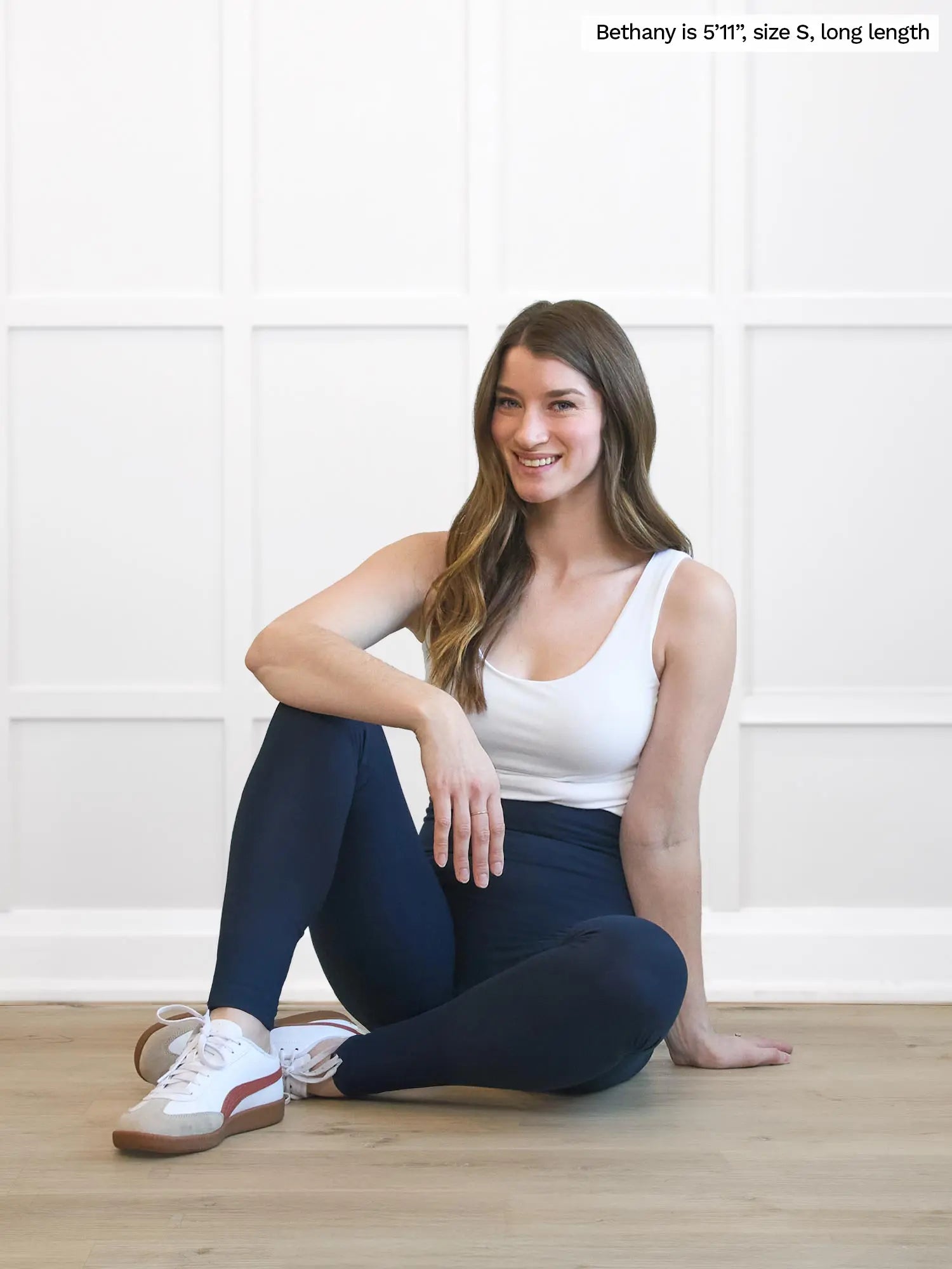 Woman sitting on the floor wearing a white tank top and dark leggings with text indicating her height and size. 