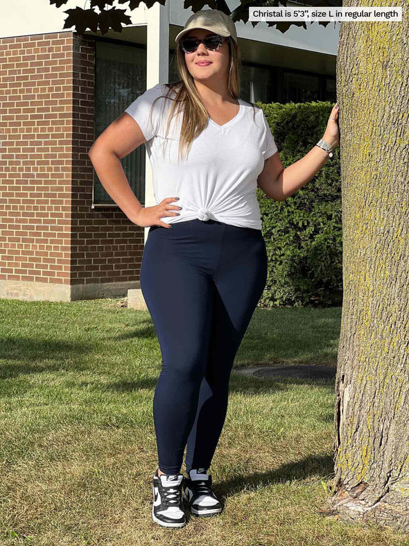 Miik model Christal (five foot three, large) standing next to a tree wearing Miik's Lisa2 high waisted legging in navy with a v-neck tee in white, sneakers, sunglasses and a cap