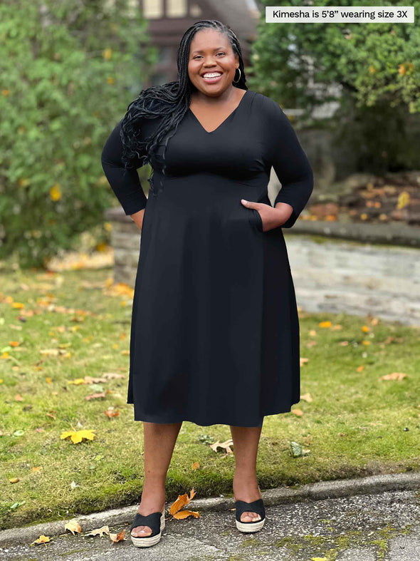 Miik model plus size Kimesha (5'8", 3x) smiling with one hand in the pockets wearing Miik's Lolly midi flounce dress with pockets in black and sandals 