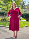 Miik model plus size Kelly (5'7", 3x) smiling, looking away with both hands on pockets wearing Miik's Lolly midi flounce dress with pockets in bordeaux