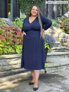 Miik model plus size Sarita (5'7", 3x) smiling while standing in front of a entry way wearing Miik's Lolly midi flounce dress with pockets in navy