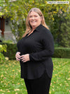 Miik model plus size Kelly (5'7", 3x) smiling while standing sideway wearing Miik's Lucia collared shirt in black along with a pant in the same colour 
