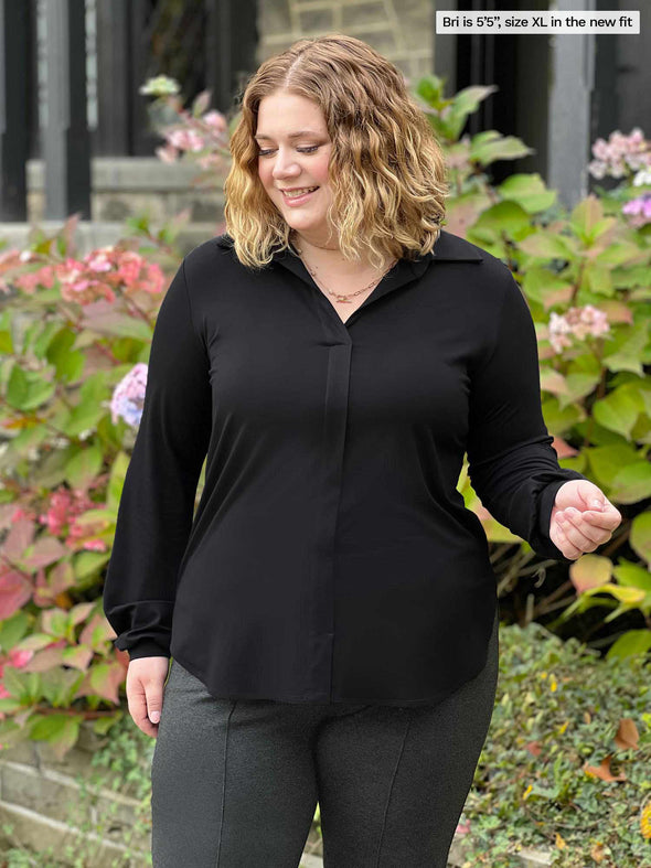 Miik model Bri (5'5", xlarge) smiling and looking down wearing Miik's Lucia collared shirt in black in the new fit along with a charcoal pant