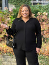Miik model Carley (5'2", xxlarge) smiling wearing an all black outfit: Miik's Lucia collared shirt, Reed pants while holding a shoulder bag 